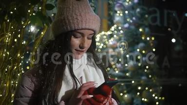 美丽迷人的女孩站在一家装饰有花环的<strong>橱窗</strong>附近，使用智能手机。 新年<strong>圣诞</strong>节